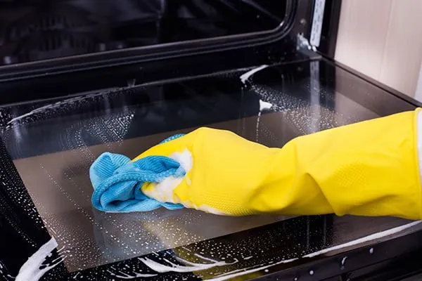 Gloved hands spreading white baking soda paste inside a greasy oven interior